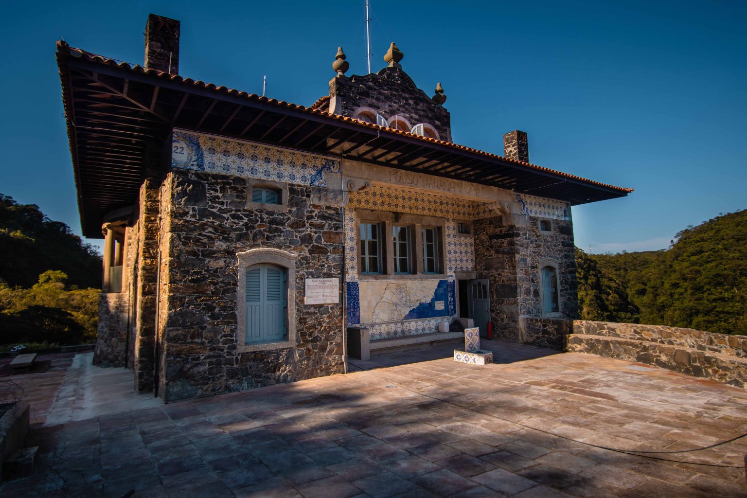 Um edifício de pedra com detalhes de azulejos fica em um terraço cercado por árvores, sob um céu limpo. Um grande mapa está em uma parede.