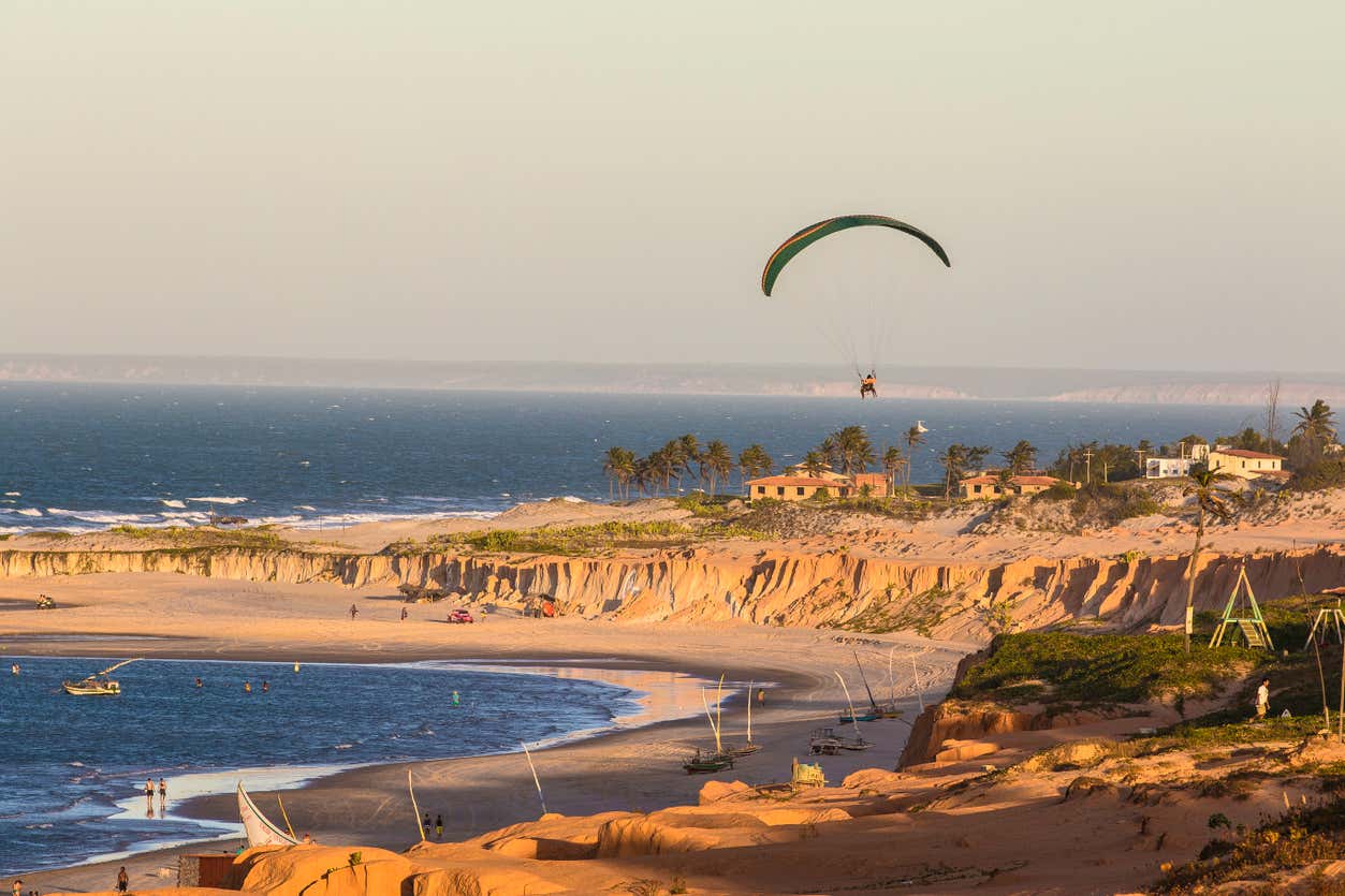 Parapente sobre uma praia arenosa com falésias, barcos na água e algumas casas e palmeiras ao longe, sob um céu limpo.