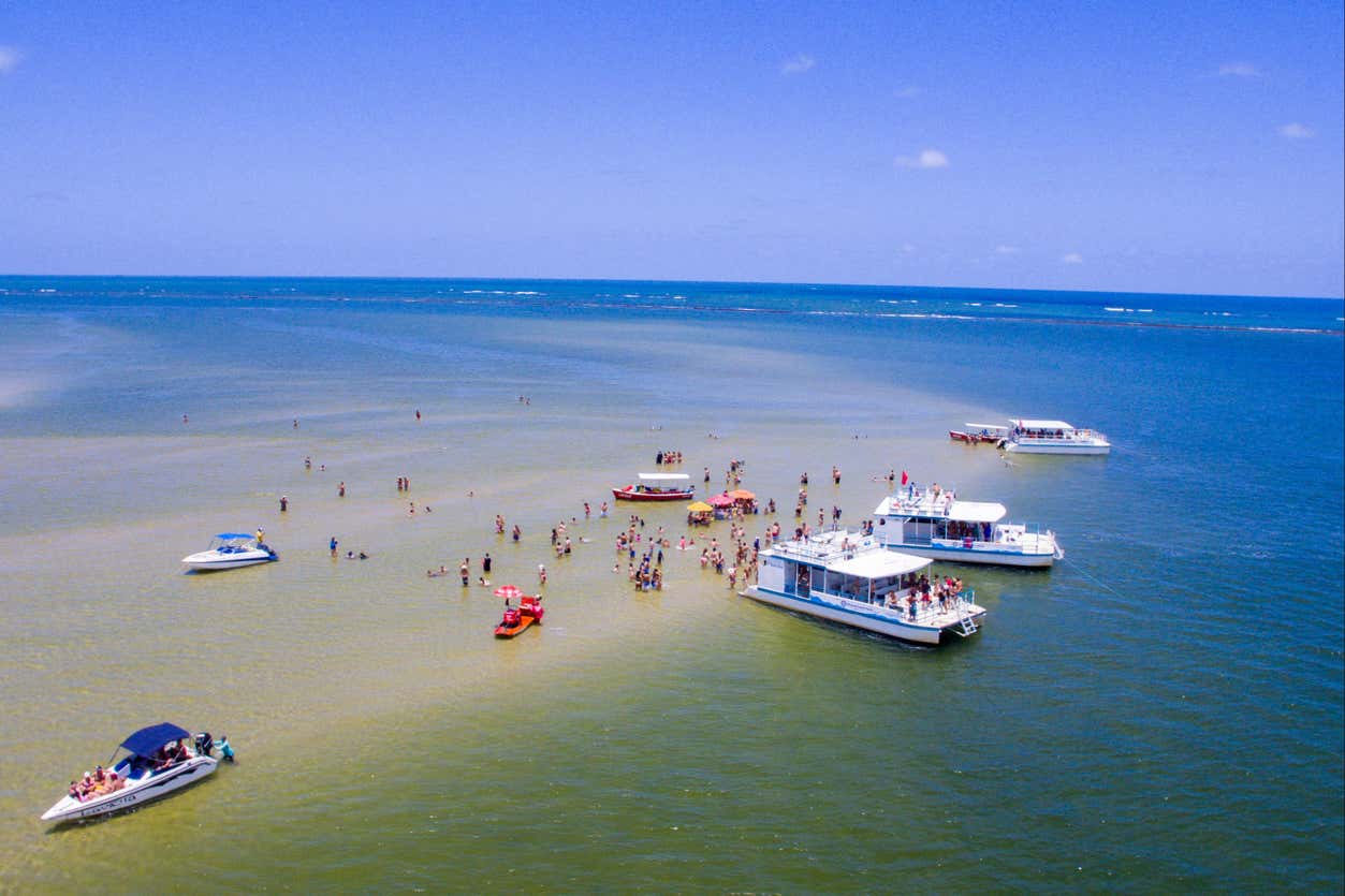 Barcos estão ancorados perto de um banco de areia com pessoas caminhando em águas rasas sob um céu azul claro.