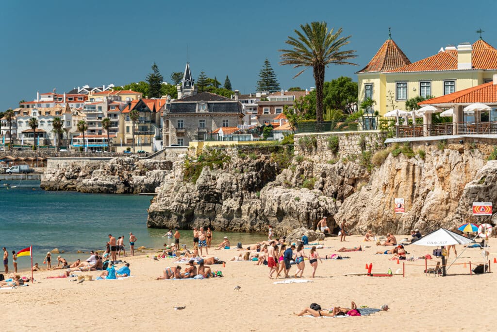 Pessoas tomando sol e nadando na Praia da Rainha em Cascais de areia com penhascos rochosos e edifícios ao fundo, sob um céu azul claro.