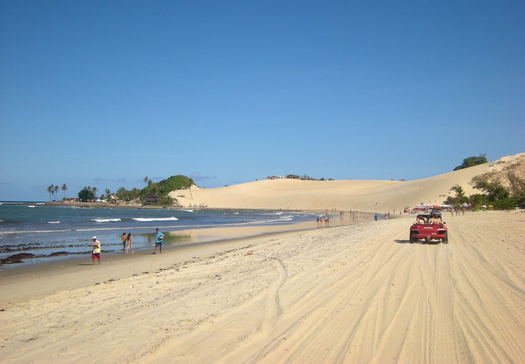 Um buggy vermelho passa por uma praia arenosa com pessoas caminhando pela costa; grandes dunas de areia são visíveis ao fundo.