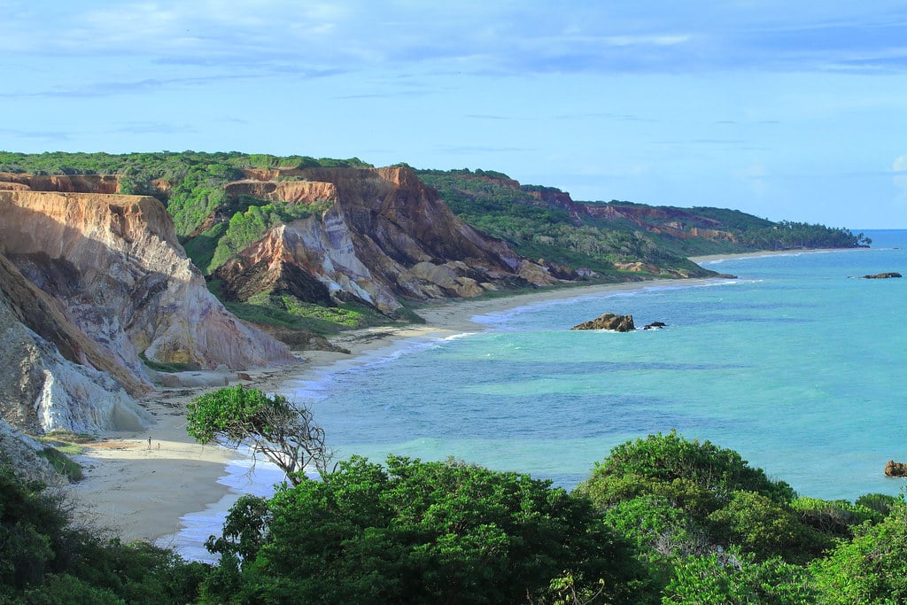 Paisagem costeira com penhascos verdes, praia arenosa e mar turquesa sob um céu parcialmente nublado. Vegetação densa em primeiro plano.
