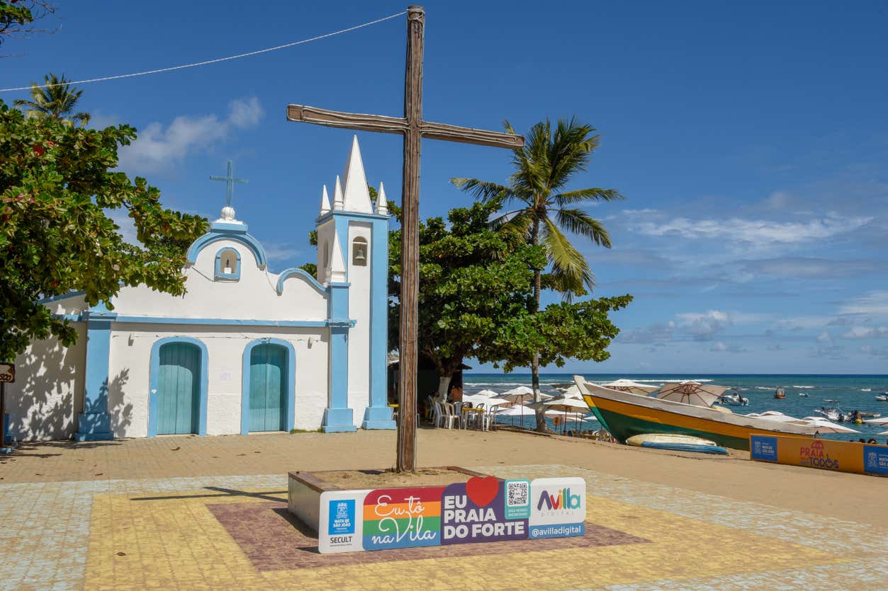 Uma capela branca e azul fica perto de uma cruz de madeira em uma praça à beira-mar com barcos e palmeiras à beira-mar.