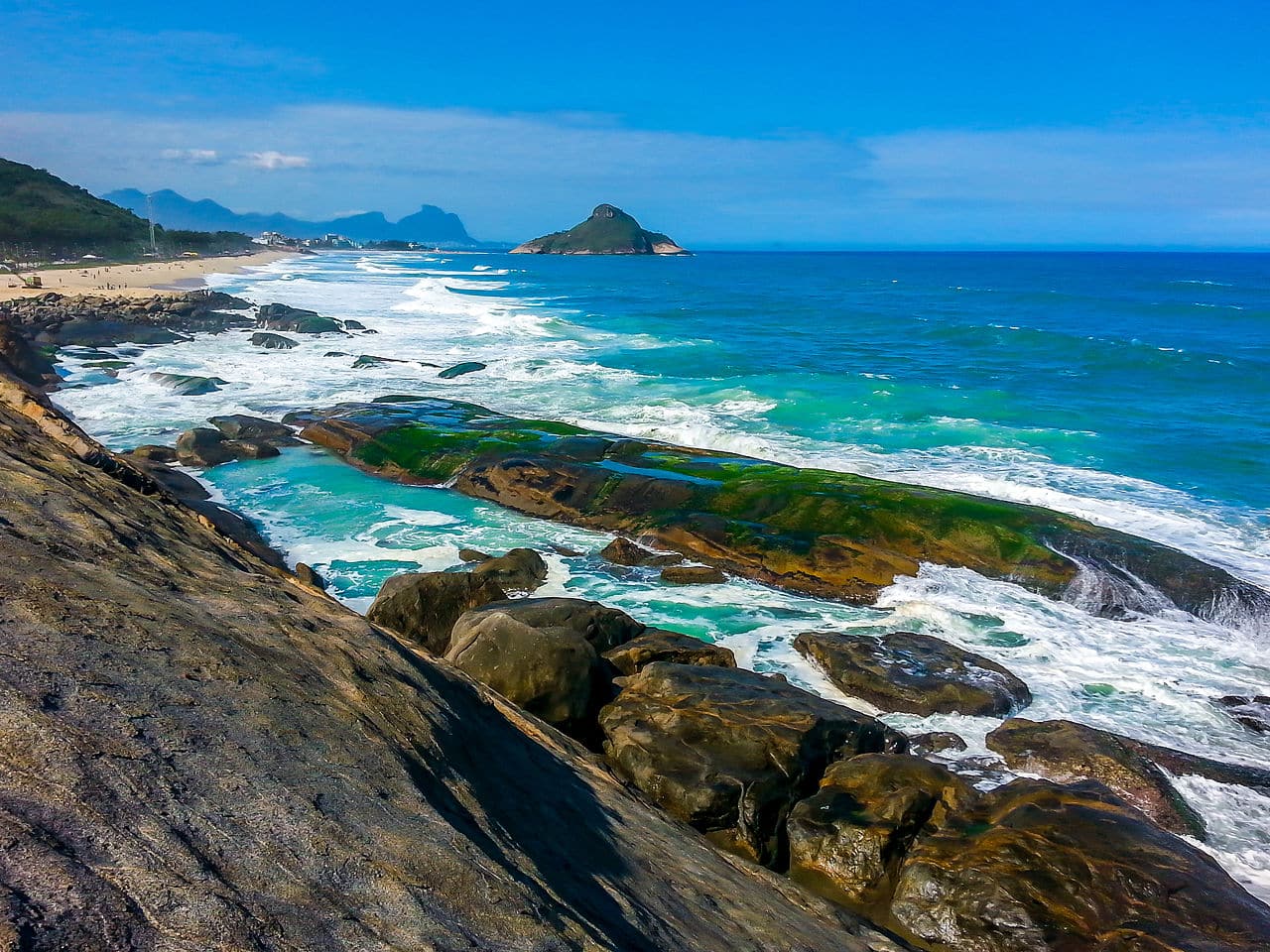 Litoral rochoso com ondas do oceano quebrando contra pedras. Praia de areia se estende para a esquerda com colinas e céu azul ao fundo. Essa é a paisagem de uma das trilhas no Rio de Janeiro.