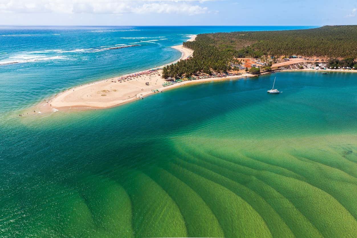 Vista aérea de uma península arenosa com uma praia, águas azul-turquesa cristalinas e um barco ancorado perto da costa, cercado por vegetação.