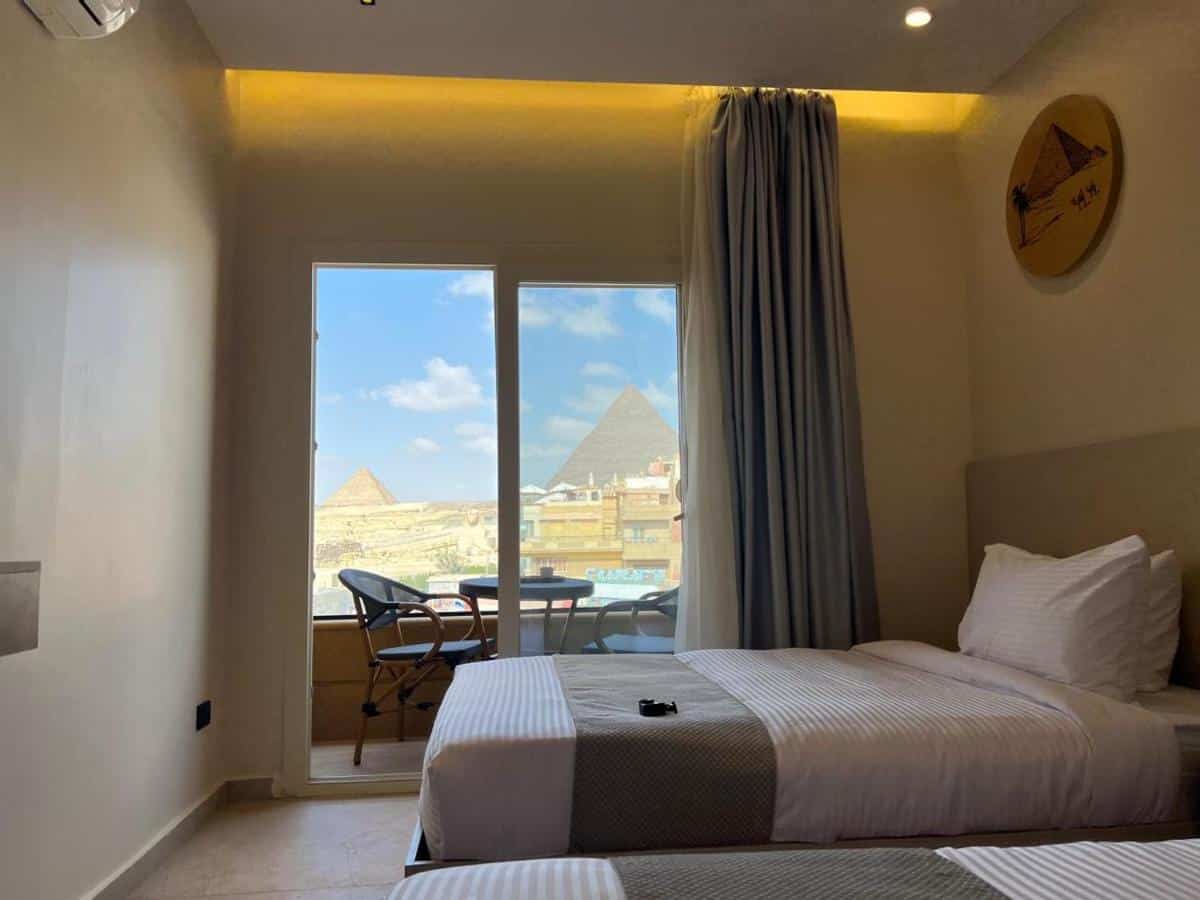 Quarto do Pyramids Island com duas camas de solteiro, varanda com cadeiras e vista para as Pirâmides de Gizé através de uma grande janela.