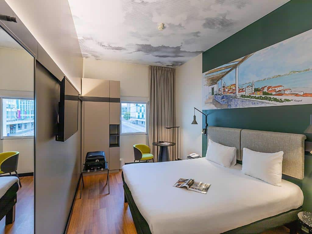 Quarto do Hotel Ibis Lisboa Parque das Nações com cama de casal, TV, duas cadeiras e um mural de parede. A luz natural entra pelas janelas à esquerda. Representa Hotéis pet friendly em Lisboa.