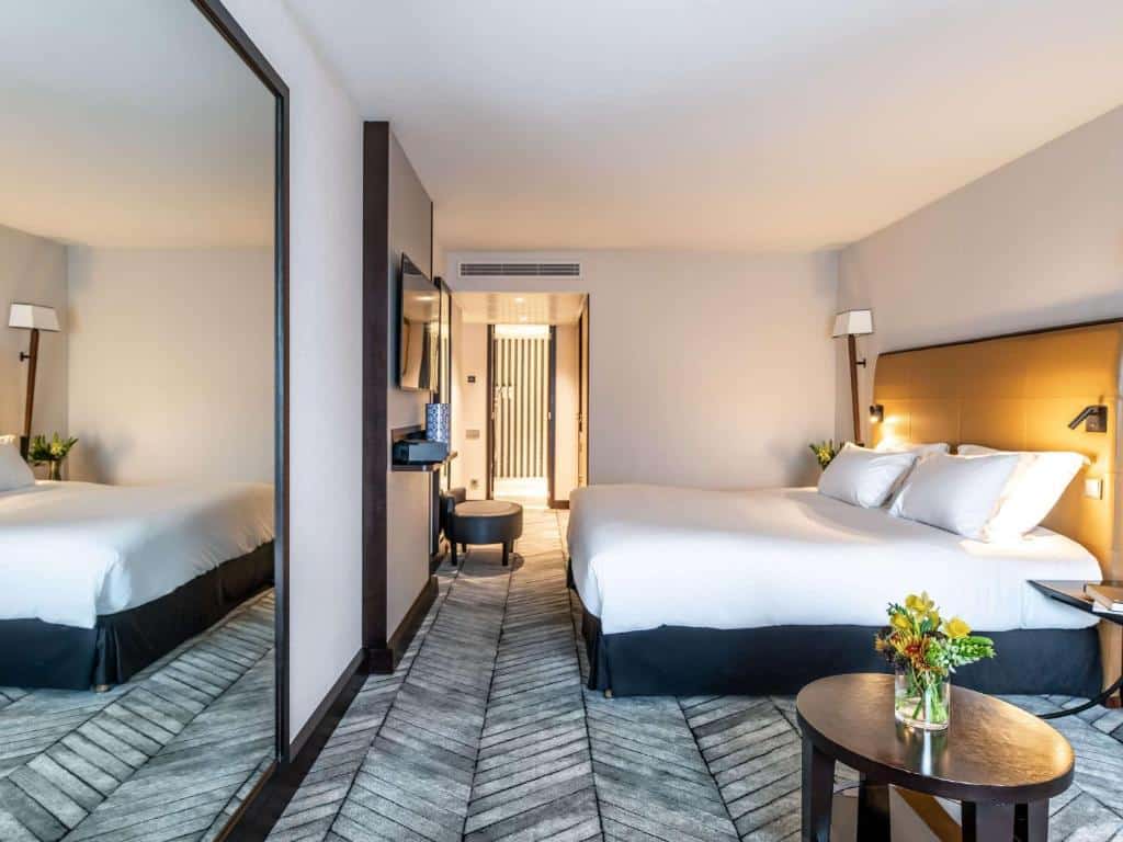 Quarto do Sofitel Lisbon Liberdade com uma cama grande, TV na parede, espelho e uma pequena mesa com flores. A porta leva a outro quarto. Representa Hotéis pet friendly em Lisboa.