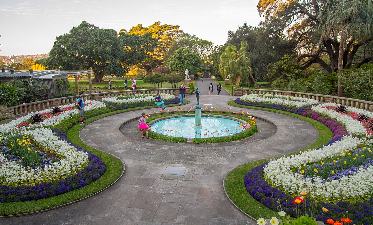 Um jardim no Royal Botanic Gardens com uma fonte circular no centro, cercado por flores coloridas, gramados bem cuidados e caminhos. Pessoas estão caminhando.