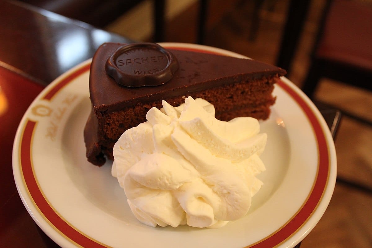 Uma fatia da Sachertorte, um bolo de chocolate com cobertura de chocolate, servida em um prato com uma colherada de chantilly.