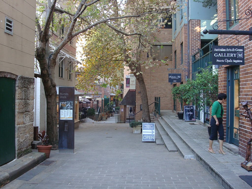 Nurses Walk no The Rock District. É um beco estreito e arborizado com prédios de tijolos, lojas e placas. Uma mulher está perto da entrada de uma galeria. Mesas são visíveis à distância. A foto ilustra o post de o que fazer em Sydney.