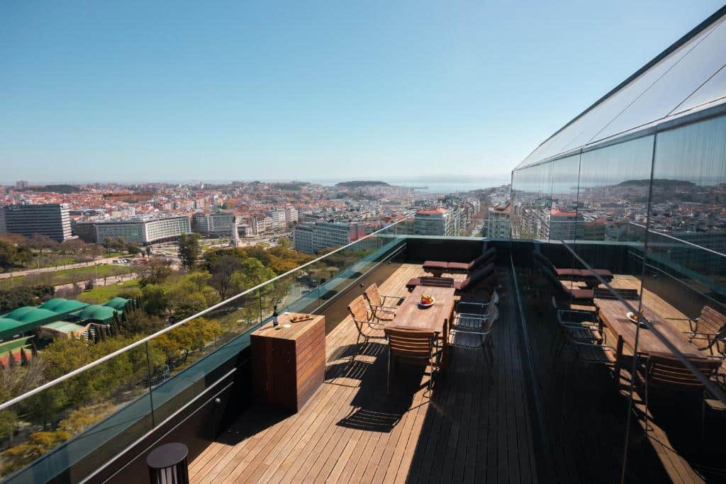 Terraço de madeira do InterContinental Lisbon by IHG com mesas e cadeiras com vista para uma paisagem urbana. O terraço tem grades de vidro, e é um dia ensolarado.