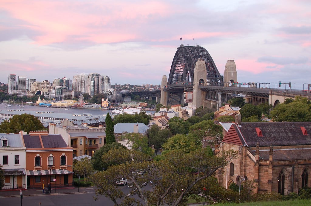 Vista da Sydney Harbour Bridge do Observatory Hill Park. É possível ver edifícios ao redor e vegetação exuberante sob um céu rosa e azul durante o pôr do sol.