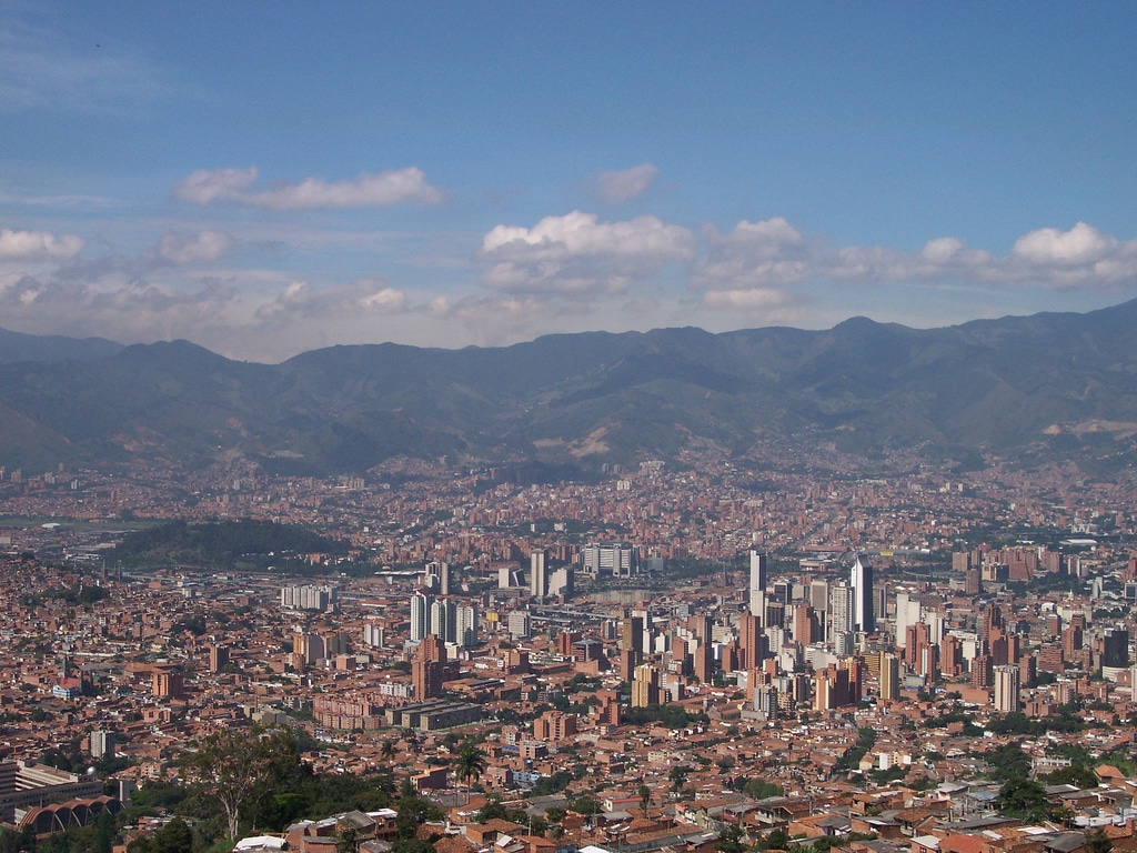 Vista panorâmica de Medellín com uma mistura de edifícios altos e áreas residenciais, tendo como pano de fundo montanhas verdes sob um céu azul. Imagem para ilustrar post sobre roteiro em Medellín.