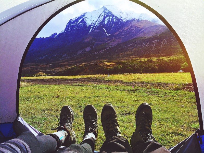 Vista de dentro de uma barraca mostrando os pés de duas pessoas em botas de caminhada, com vista para uma área gramada e montanhas cobertas de neve ao fundo.