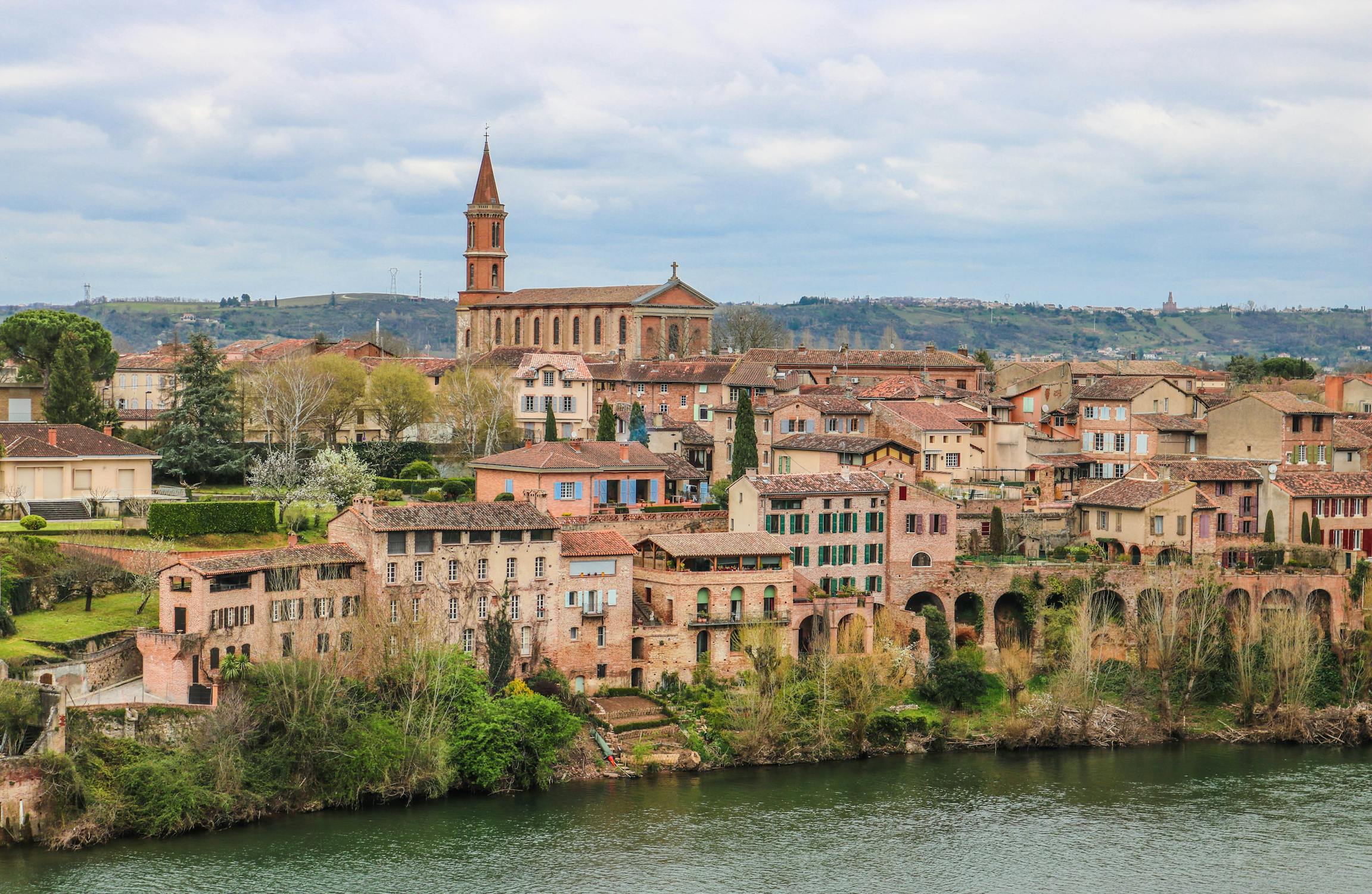 Uma vista panorâmica de uma cidade europeia com edifícios históricos, uma igreja importante com uma torre alta e um rio em primeiro plano.