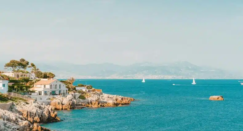 Cena costeira com penhascos rochosos, casas e veleiros em um mar azul calmo sob um céu limpo. Montanhas são visíveis à distância. Essa é a cidade de Antibes, uma das dicas de o que fazer no sul da França.