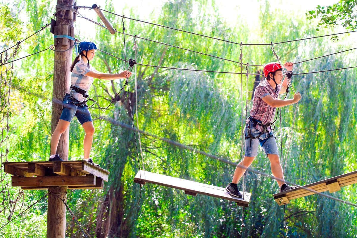 Duas crianças usando capacetes e arreios percorrem um percurso de cordas no topo das árvores, caminhando sobre pranchas de madeira entre árvores verdejantes no Parque Arví.