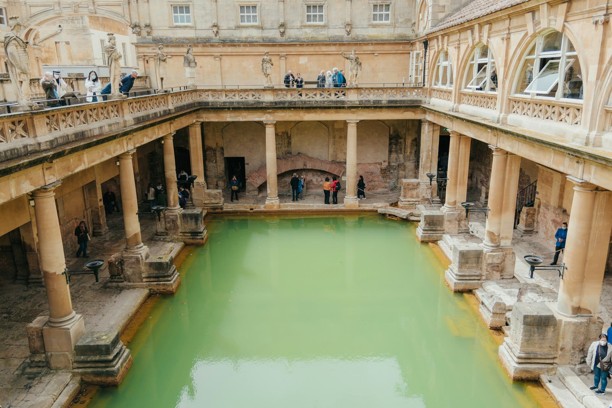 Vista dos Banhos Romanos em Bath, Inglaterra, com uma piscina verde cercada por colunas de pedra e uma passarela superior com visitantes. Ele está na lista de o que fazer em Bath.