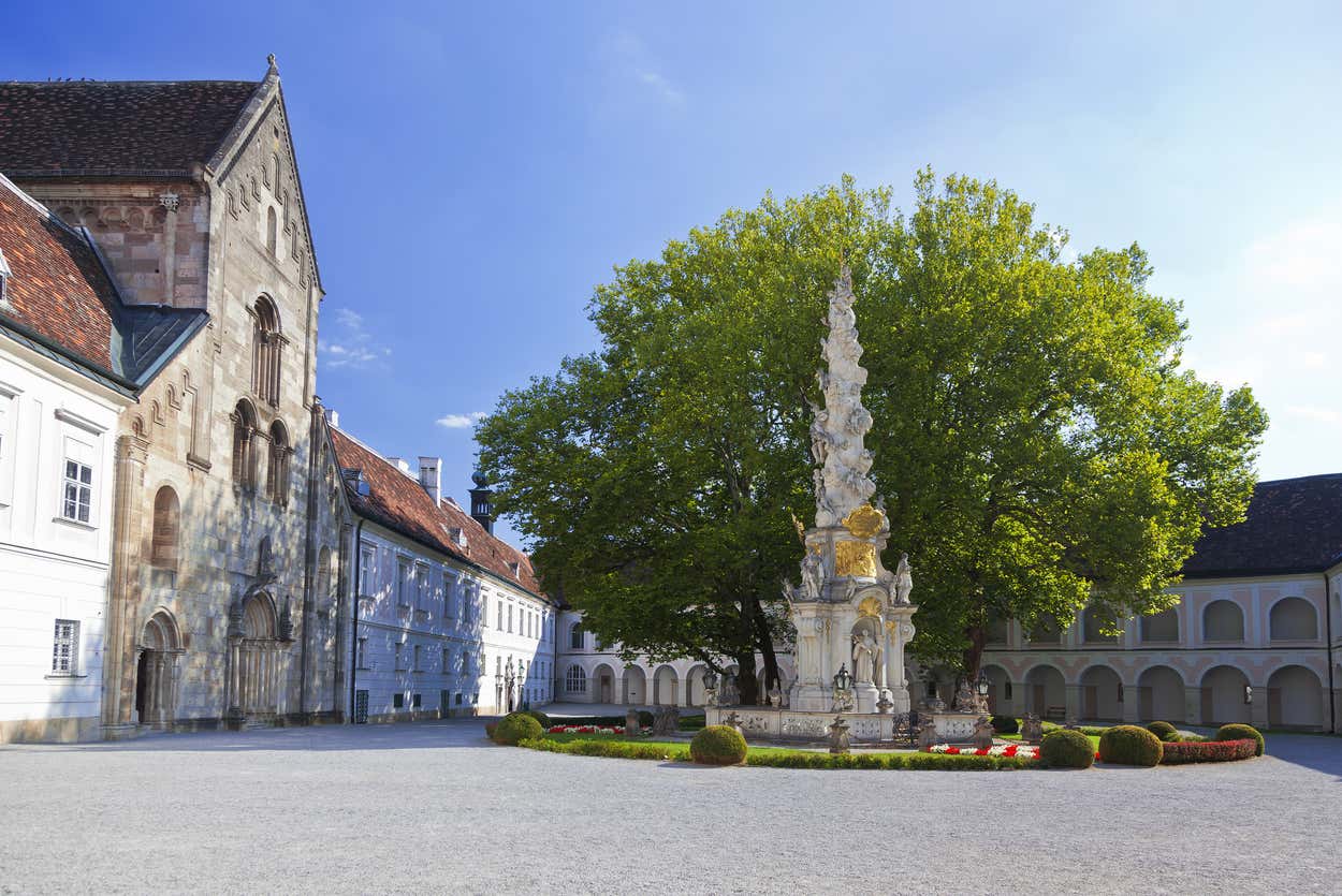 Pátio em estilo barroco com um alto monumento de pedra, cercado por edifícios históricos e uma grande árvore sob um céu azul claro. Esse é um dos pontos turísticos de Viena.