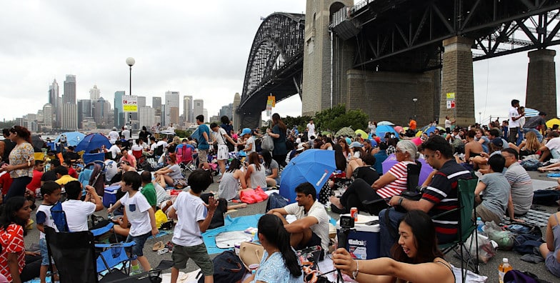 Uma grande multidão sentada com cobertores, cadeiras e guarda-sóis perto de uma ponte no Bradfield Park em um dia nublado, com o horizonte da cidade ao fundo. Ilustra o réveillon em Sydney.