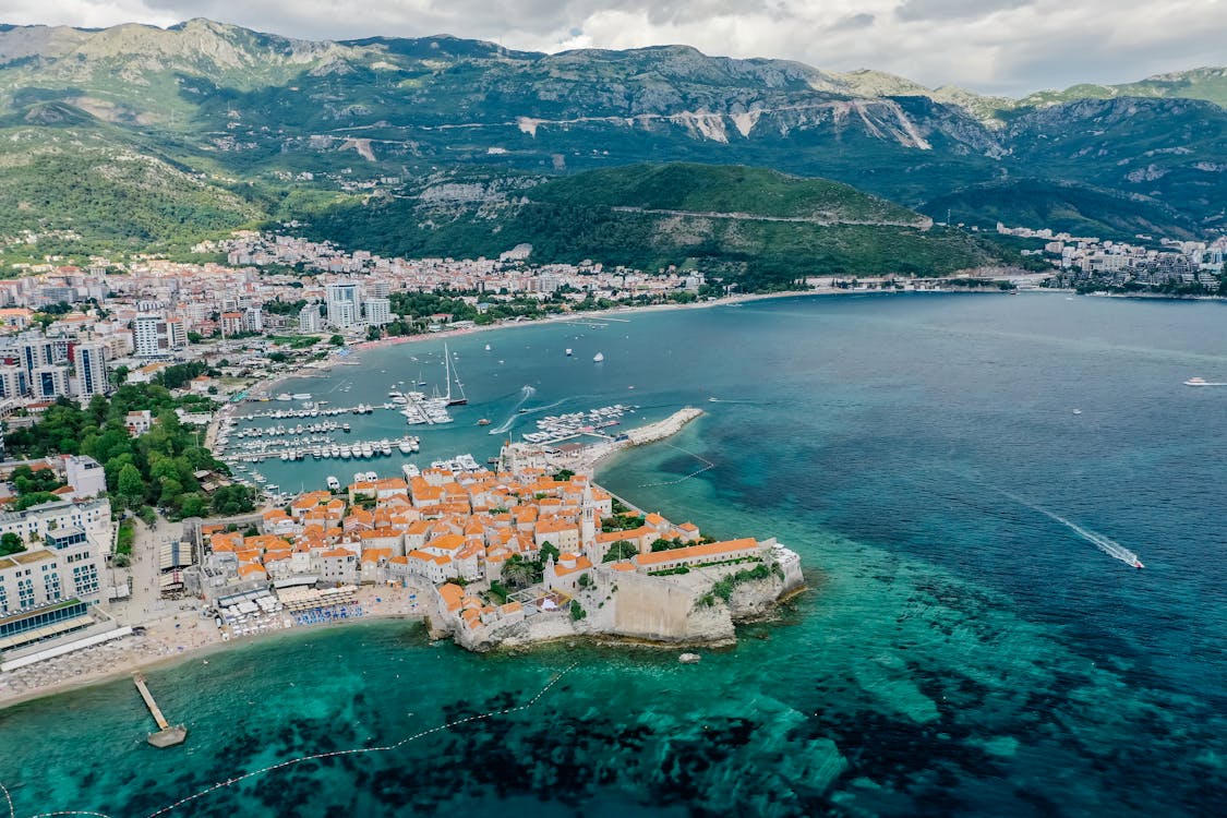 Vista aérea da cidade costeira de Budva, em Montenegro, com edifícios com telhados alaranjados, cercada por uma marina e colinas verdes sob um céu parcialmente nublado.