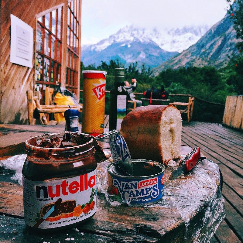 Uma mesa com Nutella, um pão, atum Van Camp's e Pringles, ao ar livre com uma montanha nevada ao fundo.