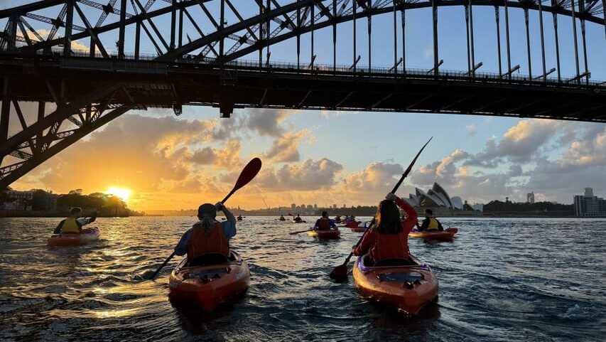 Pessoas andando de caiaque sob uma grande ponte de aço ao pôr do sol, com a Ópera de Sydney ao fundo e um céu parcialmente nublado.