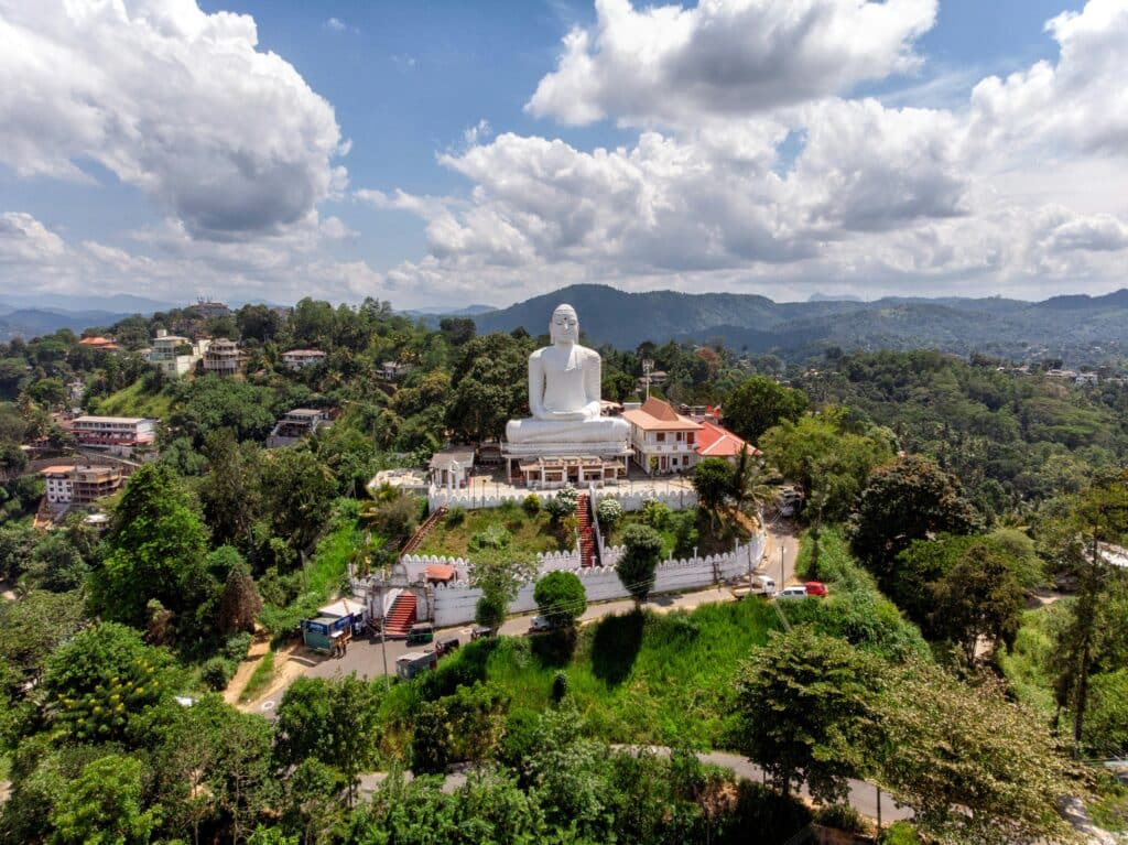 Uma grande estátua branca de Buda no topo de uma colina cercada por árvores e edifícios sob um céu parcialmente nublado, em Sri Maha Bodhi Viharaya. Representa eSIM Sri Lanka.