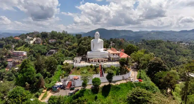 Uma grande estátua branca de Buda fica em uma colina cercada por árvores e edifícios sob um céu parcialmente nublado.