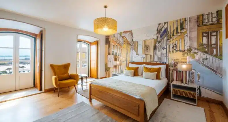 Quarto da Varandas de Lisboa com cama de madeira, cadeira amarela e um mural cênico. Duas luminárias em mesas laterais e janelas grandes com vista para fora. Representa aluguel de temporada em Lisboa.
