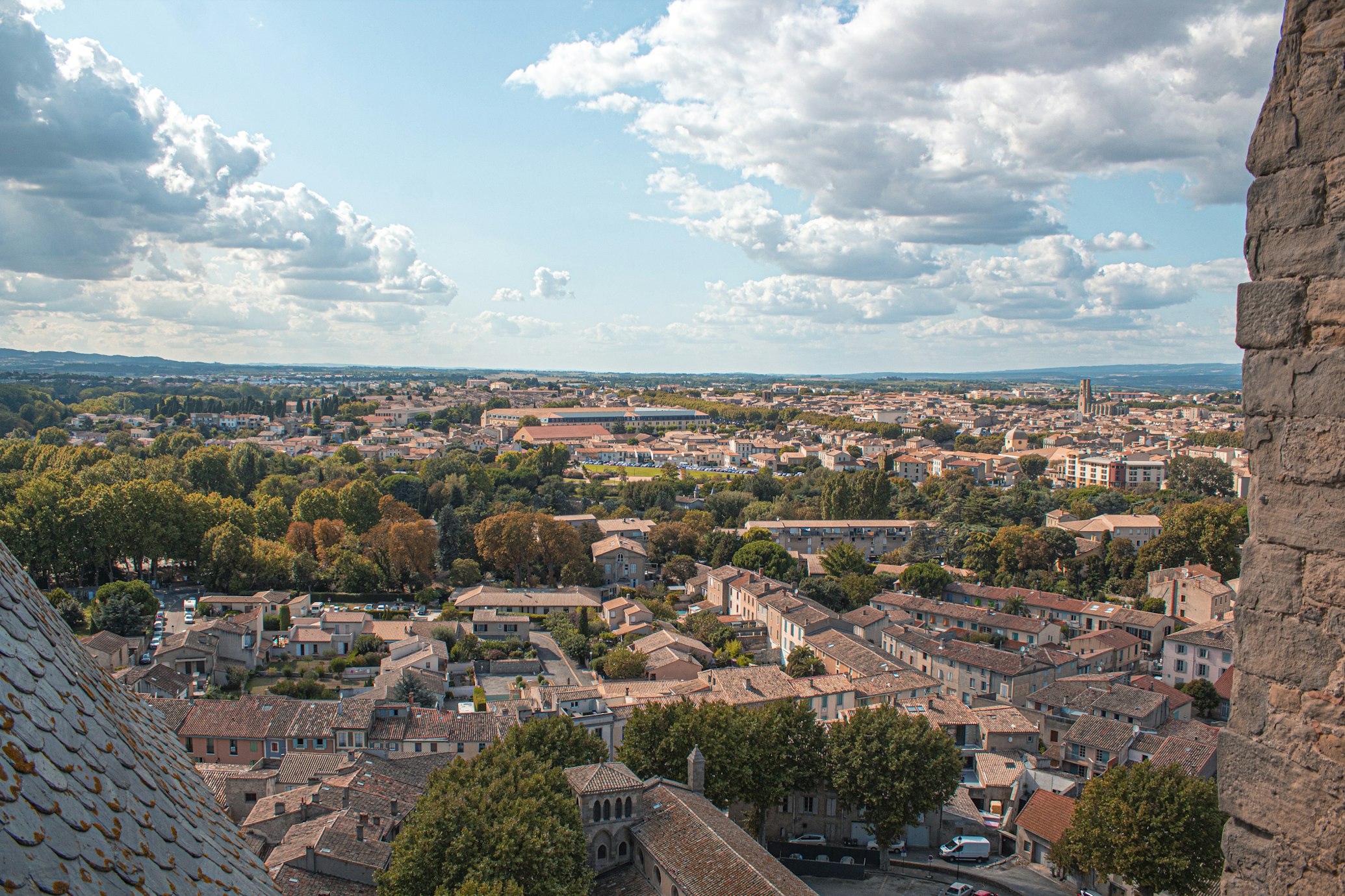 Vista aérea de uma cidade com edifícios agrupados, árvores e terras agrícolas sob um céu parcialmente nublado, vista de uma estrutura de pedra. Essa é Carcassone, uma das dicas de o que fazer no sul da França.