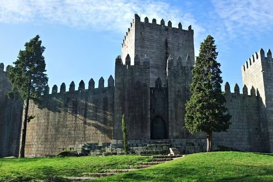Castelo de Guimarães em pedra com altas muralhas e torres ameadas, cercado por uma área gramada e árvores sob um céu azul claro.