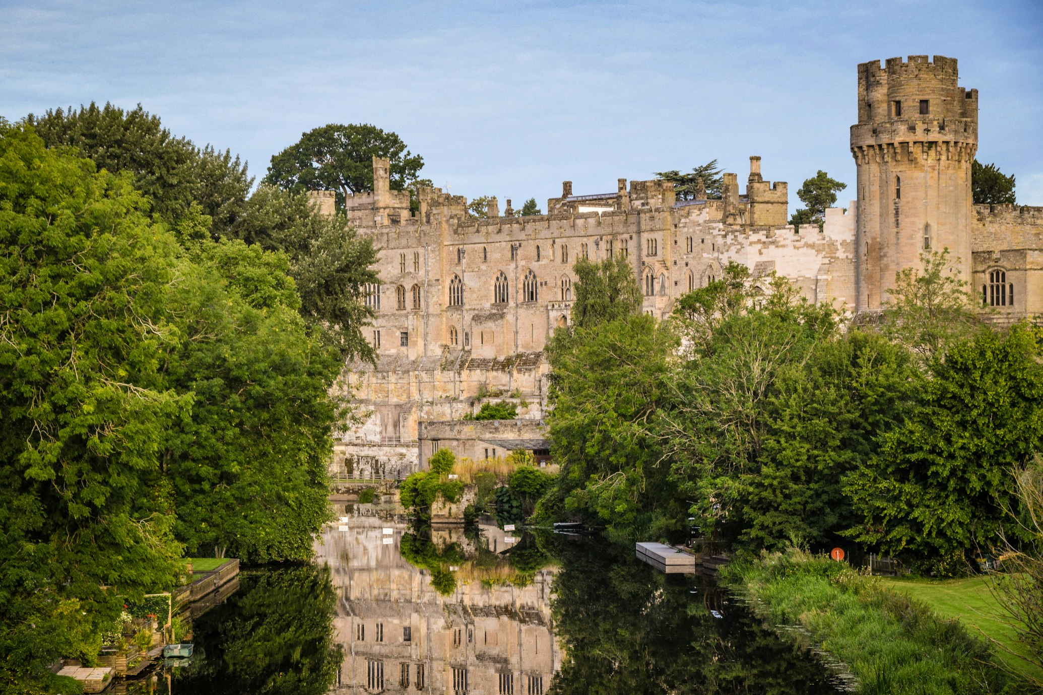 Um castelo de pedra com uma torre redonda fica atrás de uma vegetação exuberante, refletido em um rio calmo sob um céu limpo. Esse é um dos pontos turísticos da Inglaterra.