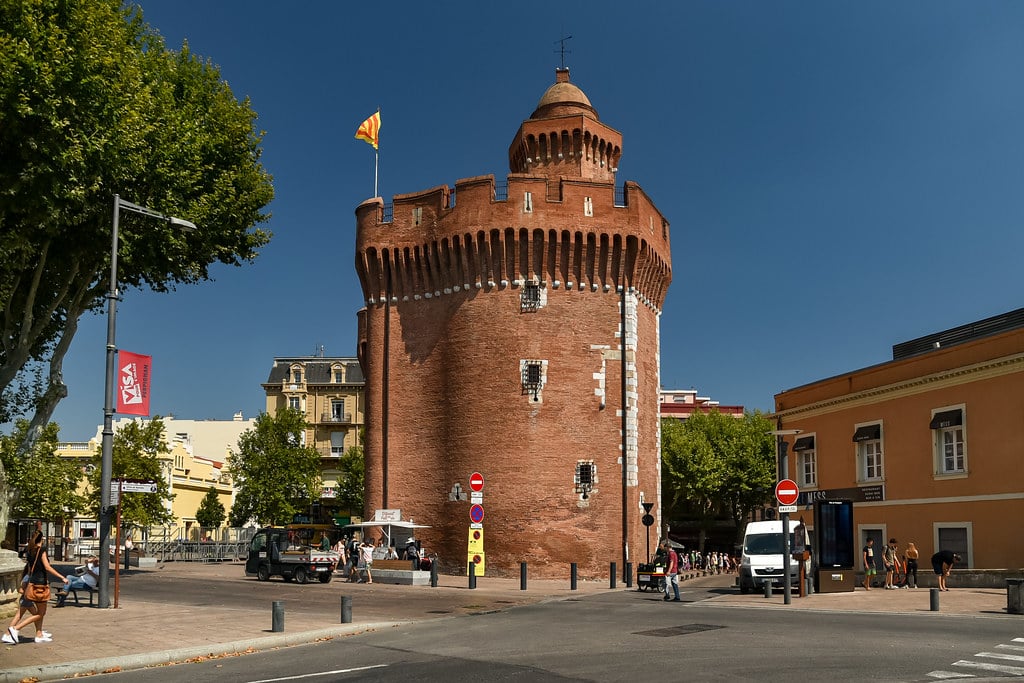 Uma torre circular de tijolos com ameias e uma bandeira no topo fica em uma praça da cidade cercada por edifícios e árvores. Essa é Perpignan, uma das dicas de o que fazer no sul da França.