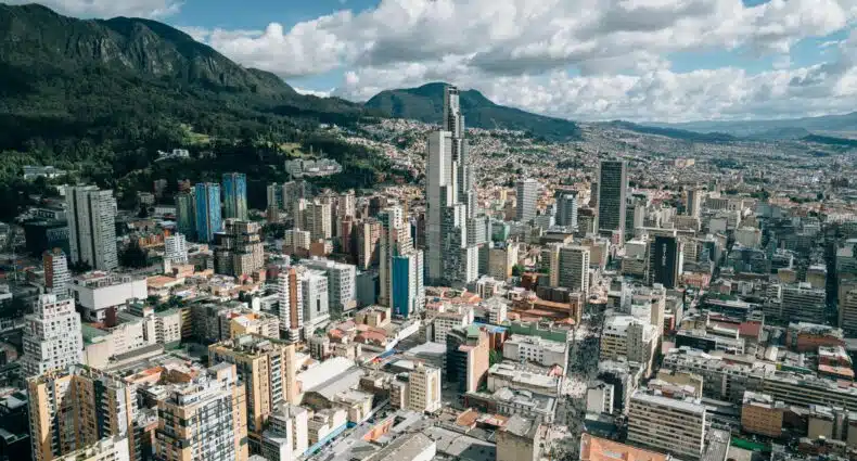 Vista aérea de uma paisagem urbana de Bogotá com edifícios altos, colinas verdes ao fundo e nuvens dispersas em um céu azul. Representa pontos turísticos de Bogotá.