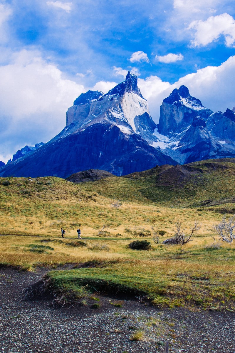 Duas pessoas caminham em uma planície gramada com montanhas recortadas e cobertas de neve sob um céu parcialmente nublado ao fundo, rumo a Torres del Paine.