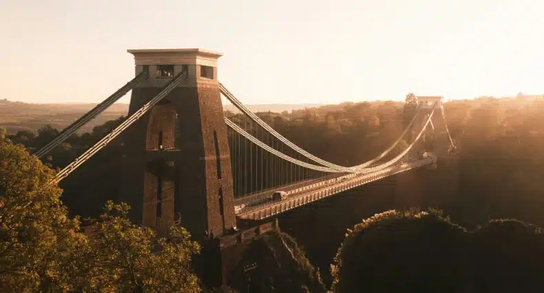 Clifton Suspension Bridge, uma ponte suspensa ao pôr do sol com árvores em primeiro plano e colinas ao fundo, criando uma atmosfera quente e dourada. A foto ilustra o post de o que fazer em Bristol.