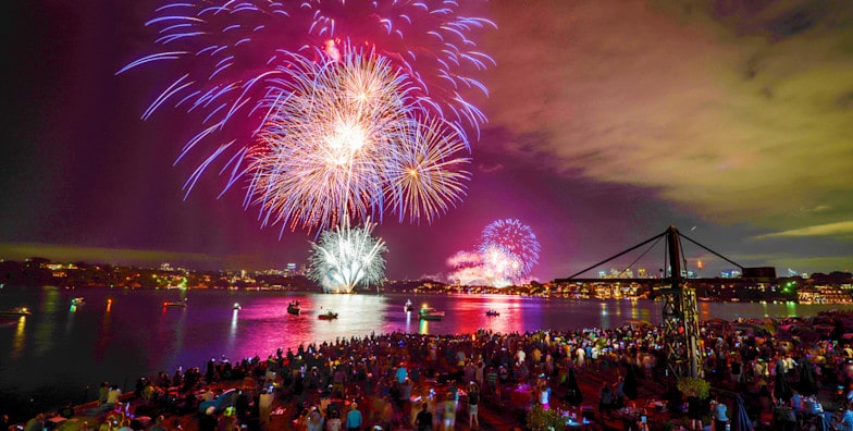 Fogos de artifício coloridos na Cockatoo Island iluminam o céu noturno sobre uma grande multidão perto da água, com luzes da cidade e barcos visíveis ao fundo. Representa o réveillon em Sydney.