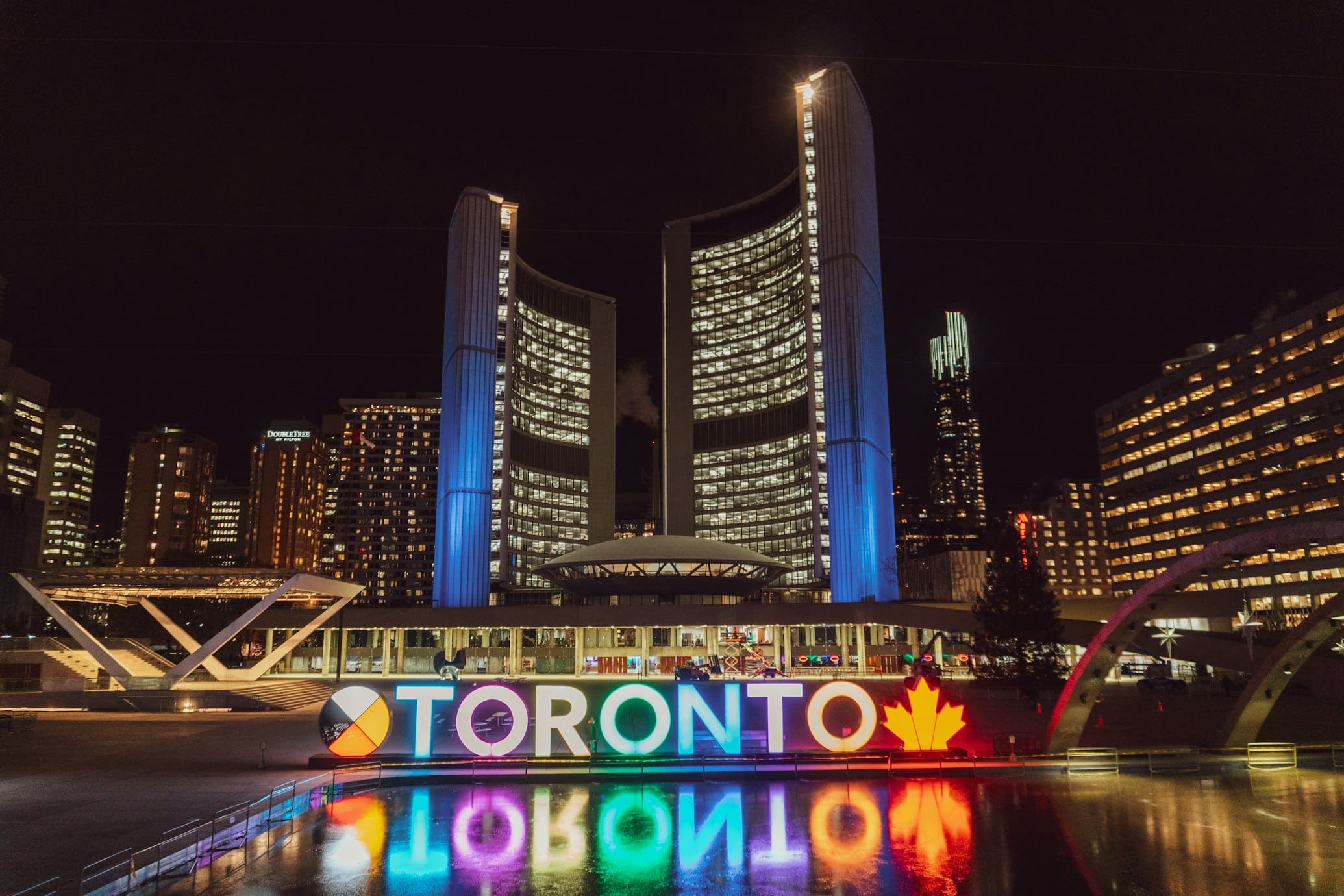 Placa de Toronto iluminada com luzes coloridas à noite, em frente a edifícios modernos iluminados, refletindo em um corpo d'água.