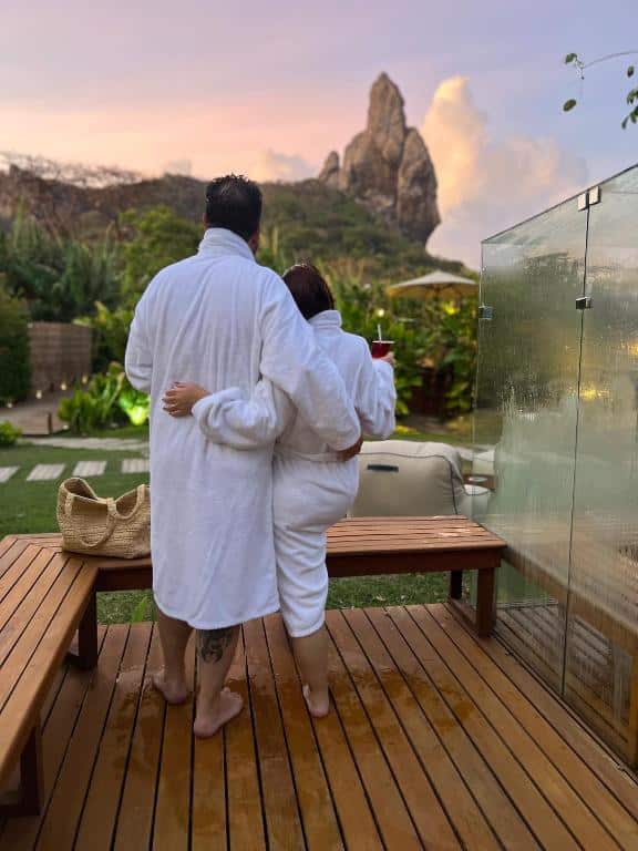 Duas pessoas em roupões brancos estão em um deck de madeira, olhando para colinas rochosas distantes. Uma segura uma bebida; uma bolsa repousa perto.