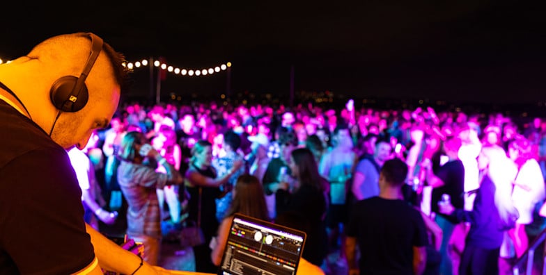DJ usando fones de ouvido toca música em uma festa ao ar livre à noite, com uma grande multidão dançando sob luzes coloridas no Dudley Page Reserve, no réveillon em Sydney.
