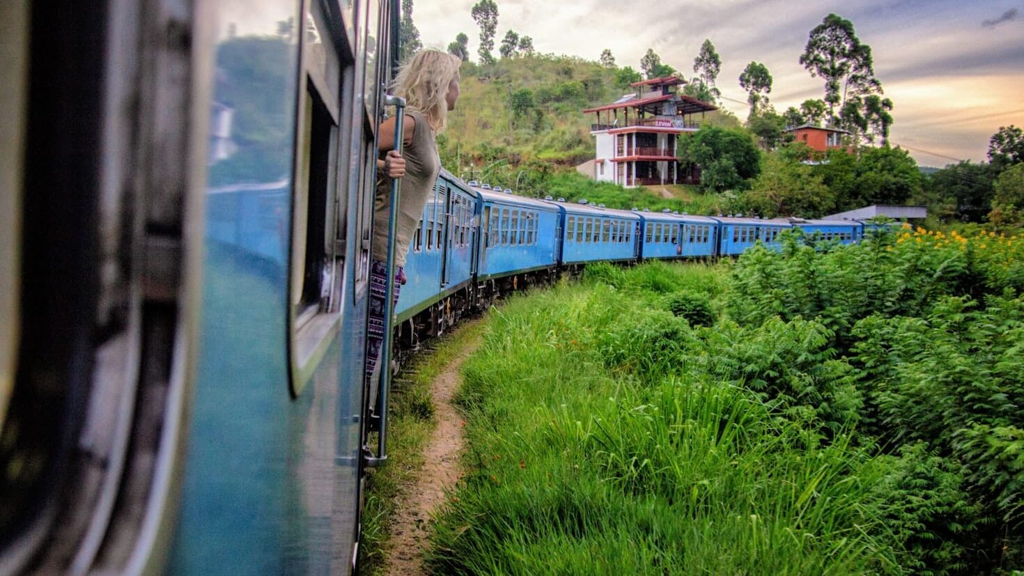 Uma pessoa se inclina para fora de um trem azul na Ponte dos Nove Arcos em movimento passando por uma paisagem verdejante com colinas e uma casa ao fundo.