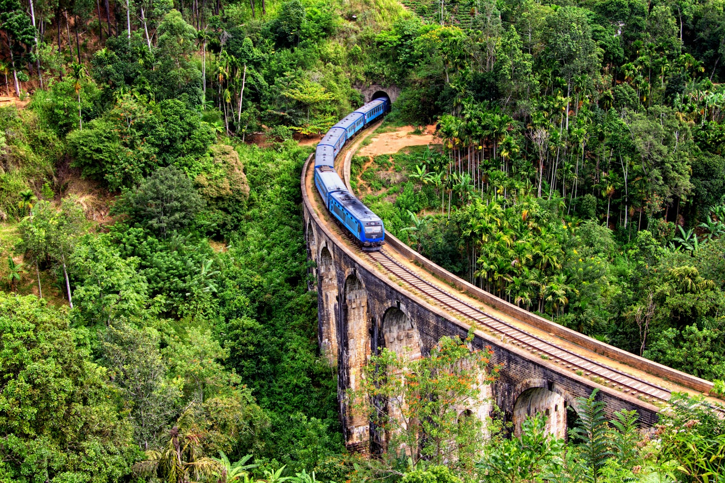 Um trem azul em Ella passa por uma ponte de pedra curva cercada por densa vegetação verde e árvores. Representa o que fazer no Sri Lanka.