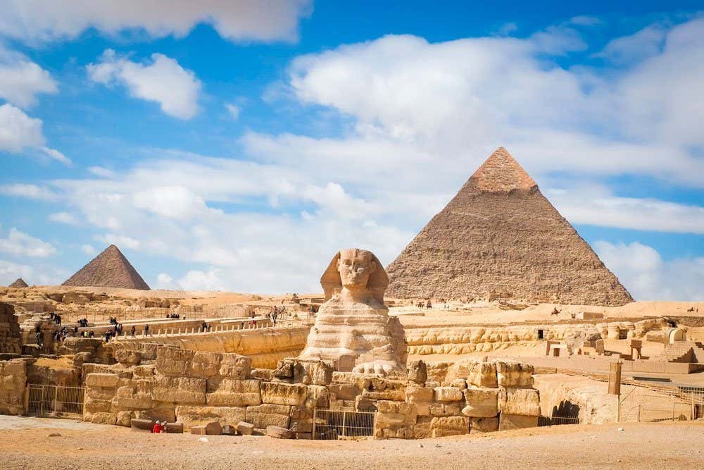 A Grande Esfinge e duas pirâmides no Cairo, sob um céu parcialmente nublado em uma paisagem desértica com turistas dispersos.