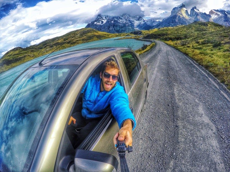 Pessoa de jaqueta azul se debruça para fora da janela do carro, tirando uma selfie com montanhas e um céu limpo ao fundo.