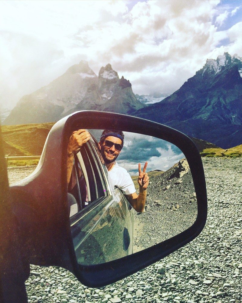 Uma pessoa de óculos escuros se inclina para fora da janela de um carro mostrando um sinal de paz, refletido no espelho lateral. Torres del Paine ao fundo.