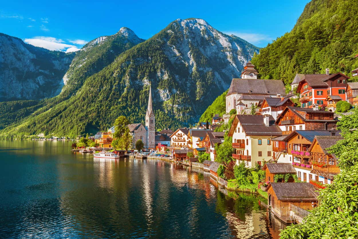 Vista panorâmica de uma vila à beira do lago com casas coloridas, uma igreja e montanhas sob um céu azul claro em Hallstatt, Áustria.