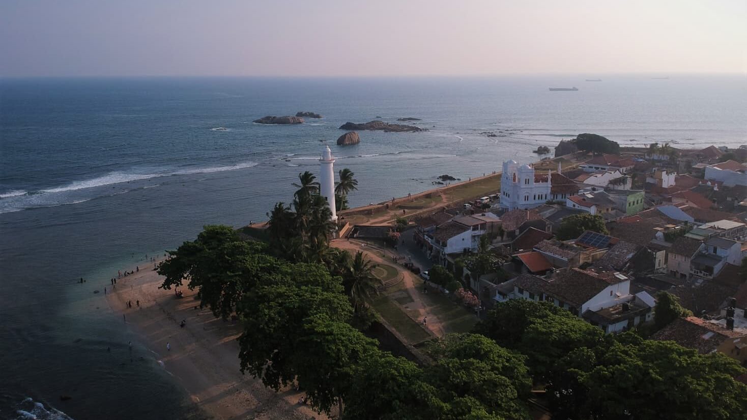 Vista aérea em Galle com um farol, árvores verdes e casas à beira-mar sob um céu limpo. Representa o que fazer no Sri Lanka.