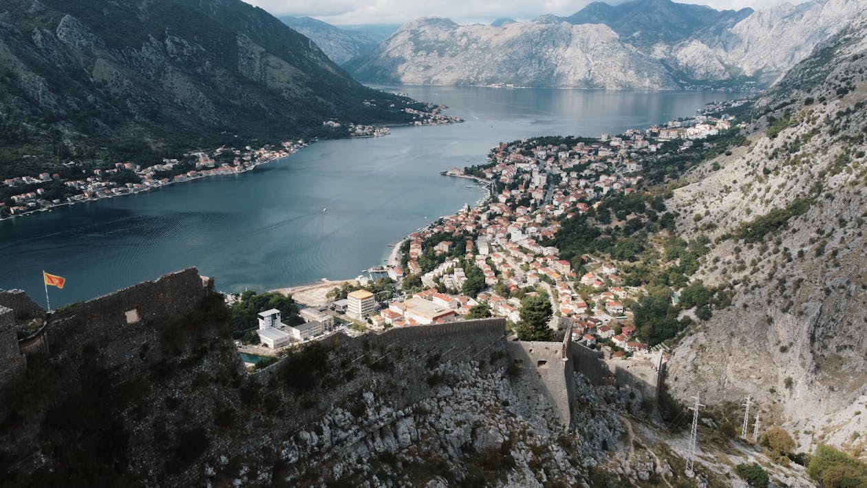 Vista aérea da cidade costeira de Kotor, em Montenegro, com edifícios de telhados vermelhos, cercada por montanhas e um grande corpo de água, sob um céu nublado.